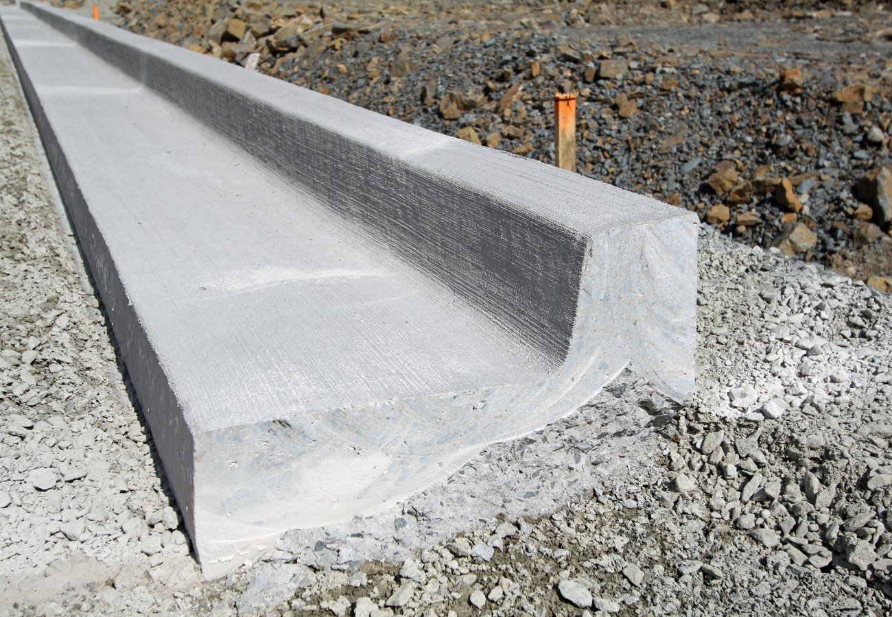 A close-up view of a freshly constructed concrete curb on a gravel roadbed in Boynton Beach. The curb has a clean, smooth surface with a slight bevel on the edge. Orange markers are seen in the background around the construction site, likely managed by a stamped concrete contractor from Palm Beach County.