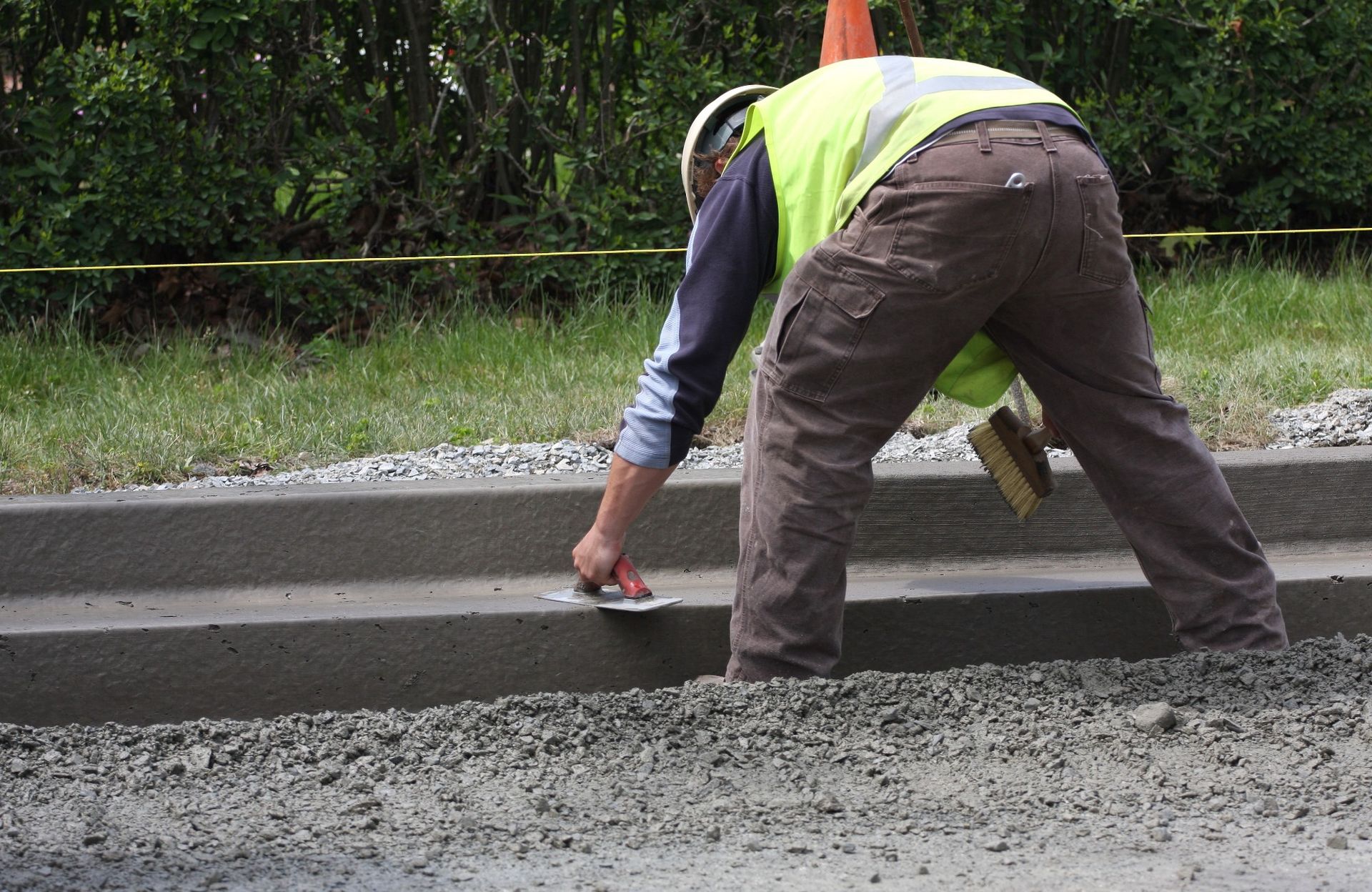 A licensed concrete contractor wearing a reflective vest and hard hat smooths out wet concrete on a step with a hand trowel. Gravel and grass are visible in the background, with a yellow string line marking the construction area.