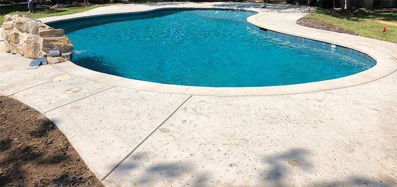 An irregularly shaped outdoor swimming pool with clear blue water is surrounded by a light-colored, slightly speckled Palm Beach Stamped Concrete deck. There is a rock formation to the left and grass with some patches of bare ground nearby. Shadows of trees are cast on the concrete in West Palm Beach, FL.