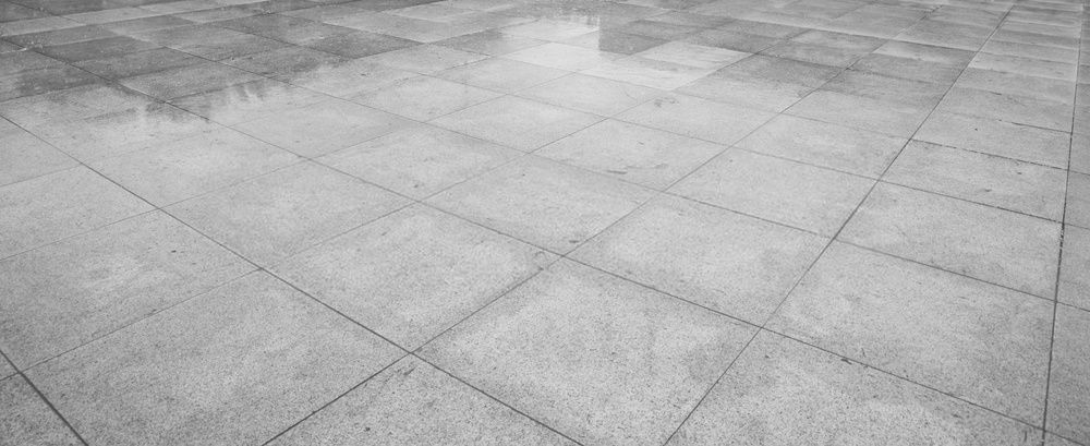 A monochrome image of a wet, empty tiled pavement with a few puddles scattered across the surface. The tiles are neatly aligned in a geometric pattern, reflecting subtle shades of grey, reminiscent of the meticulous work by concrete contractors in Palm Beach County.