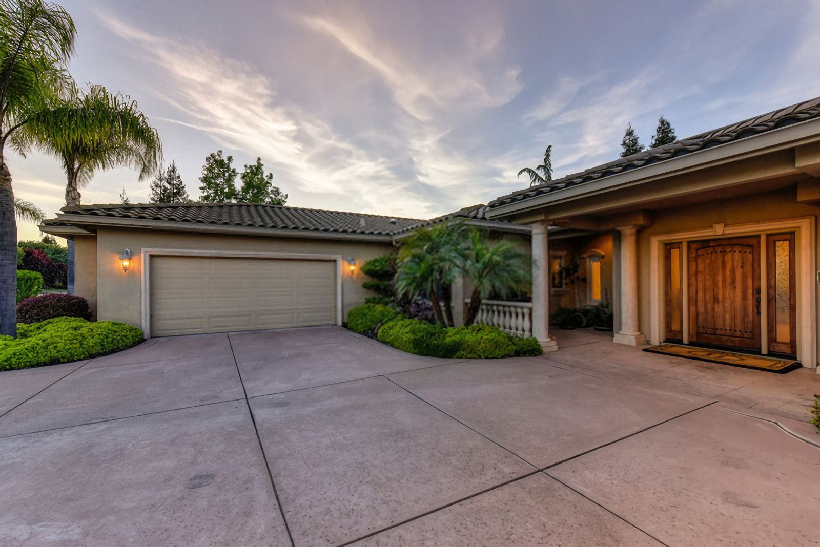 A suburban house with a decorative tile roof, featuring a double garage on the left and a wooden front door to the right under a covered porch. The wide driveway showcases Palm Beach Stamped Concrete, and the landscaped yard includes trees, shrubs, and a palm tree. The sky is partly cloudy during sunset.