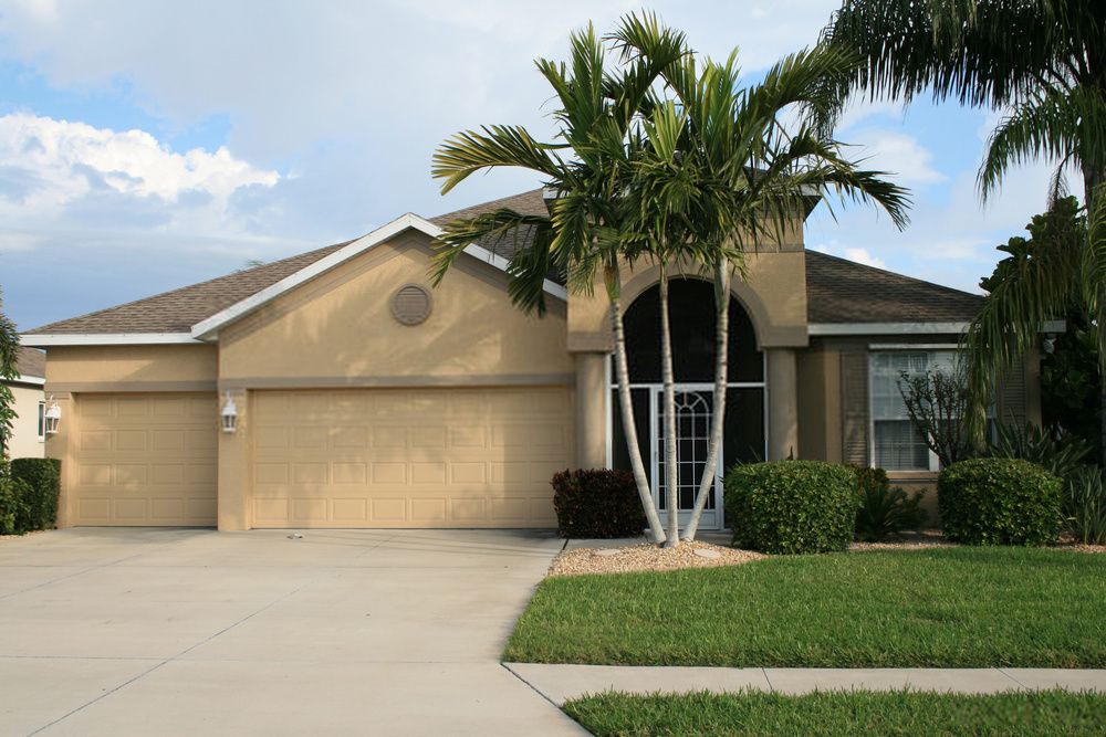 A single-story beige house with a two-car garage, surrounded by manicured bushes and palm trees, is set against a partly cloudy sky. A neatly crafted Palm Beach Stamped Concrete driveway leads to the garage, while a lawn area with green grass and a small garden adorns the front.