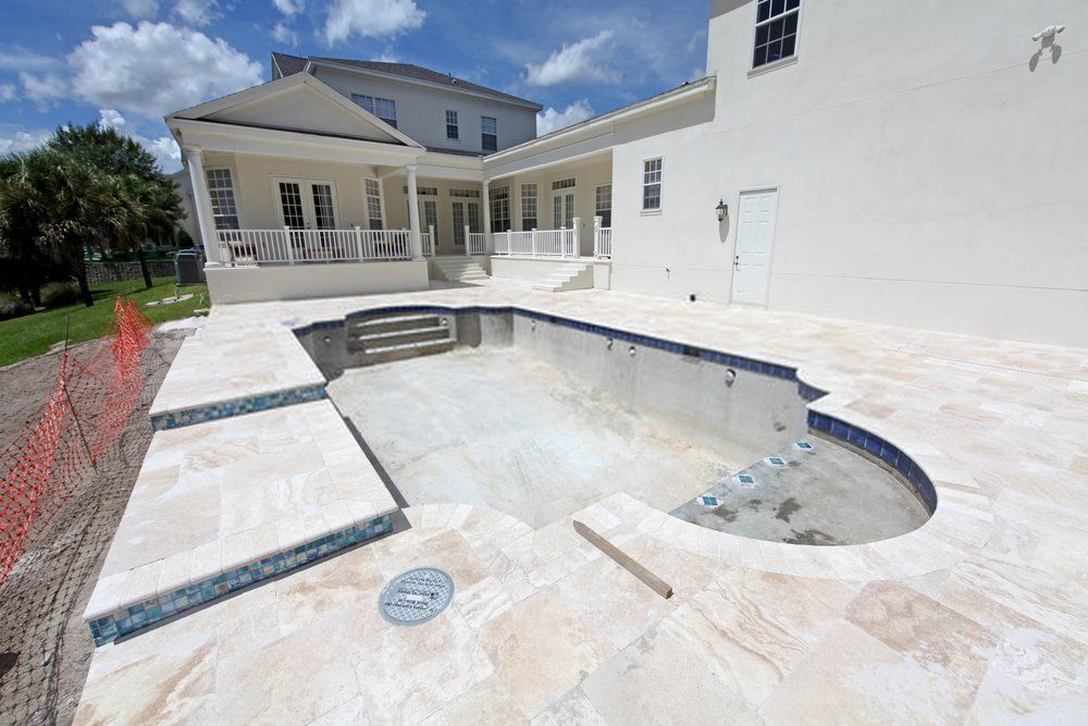An empty, unfinished swimming pool in a large backyard hints at the recent work of concrete contractors in West Palm Beach, FL. The pool is surrounded by beige tiles, and an orange construction fence is partially visible on the left. A large, two-story white house with a porch and multiple windows stands proudly in the background.