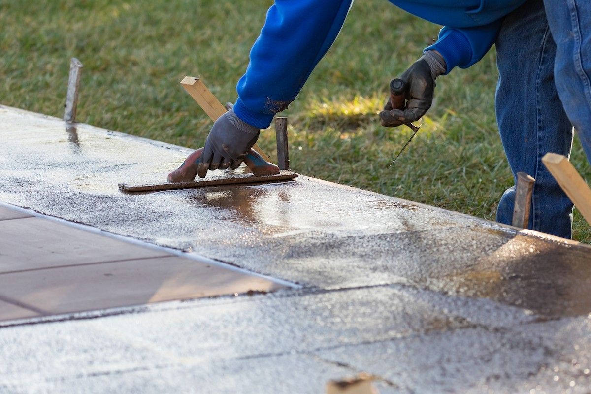 A person in a blue sweatshirt and jeans smooths out wet concrete with a trowel on a rectangular surface. Wooden stakes and a grassy area can be seen in the background, as they work on Palm Beach Stamped Concrete.