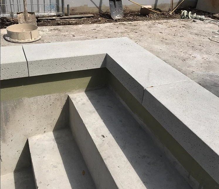 A close-up view of a partly constructed concrete staircase with three steps and an L-shaped corner. The construction appears unfinished, with building materials and a fence visible in the background. A trowel and other construction tools are scattered around, indicative of a decorative concrete installation in progress.