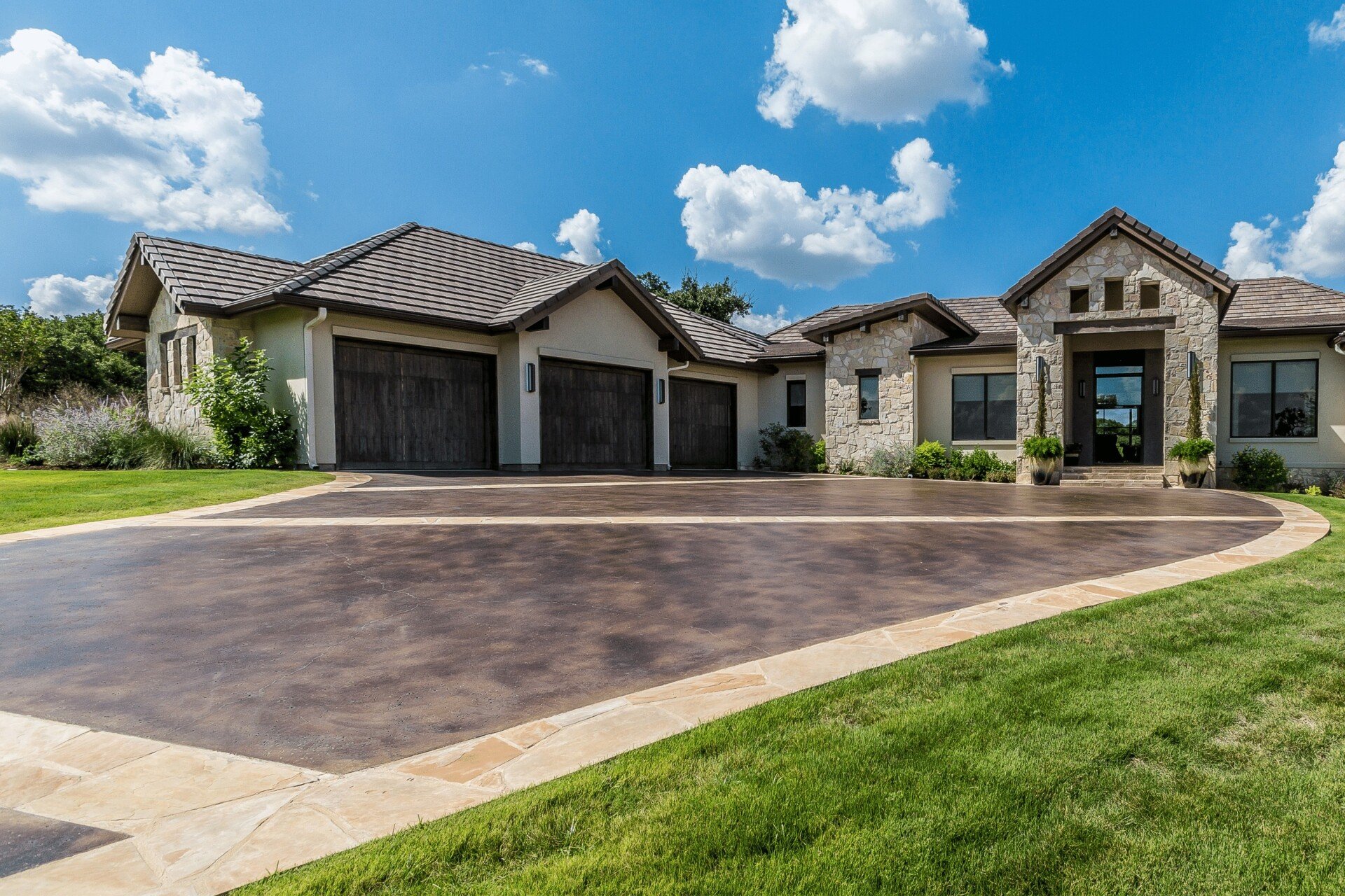 A modern, single-story house with a stone facade and multiple gables stands proudly in Palm Beach County. The property features a spacious driveway crafted by a stamped concrete contractor, leading to a three-car garage. Set against a clear blue sky with fluffy white clouds, the house is surrounded by well-maintained green lawn.