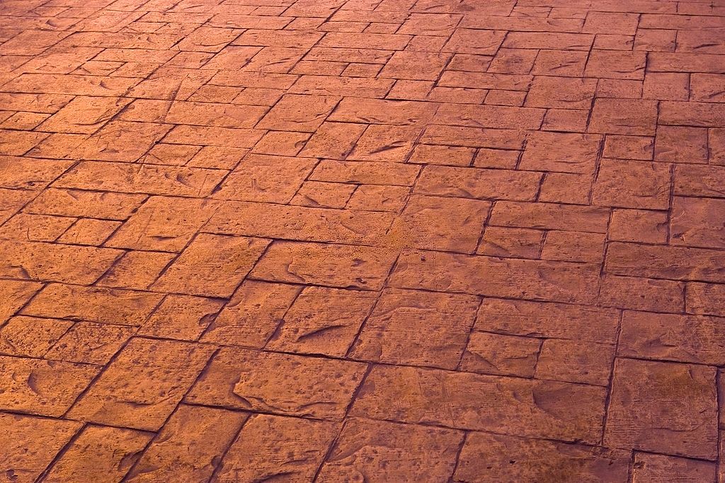 A close-up view of a ground surface with a patterned design made of textured, rectangular stone tiles in varying shades of warm brown. The sunlight accentuates the texture and color variations, creating a visually appealing effect that showcases the expertise of concrete contractors in Palm Beach County.
