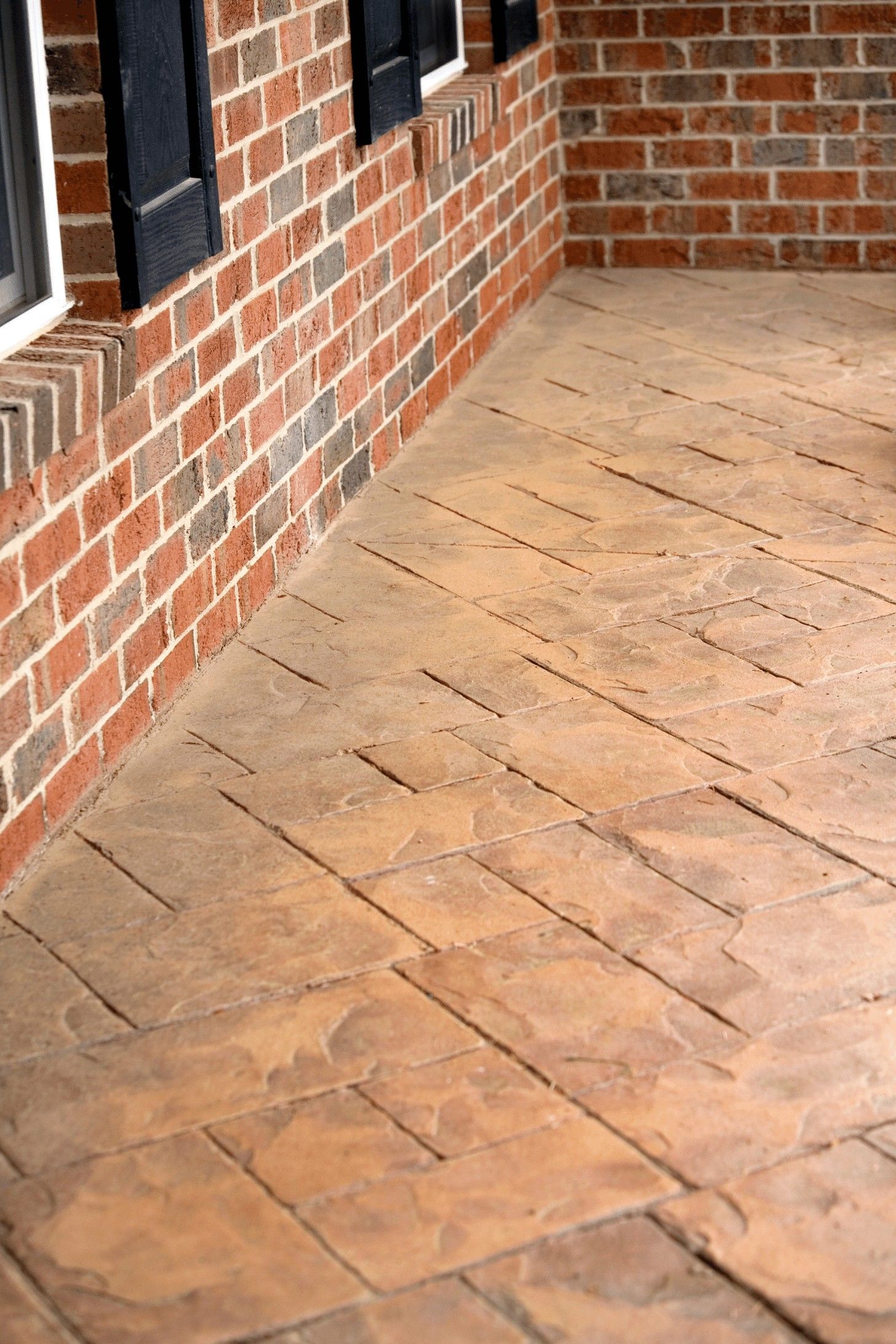 A close-up view of a building's exterior in Juno Beach showcasing a brick wall with a few windows having dark blue shutters and a stamped concrete patio by Palm Beach Stamped Concrete. The patio features terracotta-colored tiles arranged in a geometric pattern, highlighting their decorative concrete services.