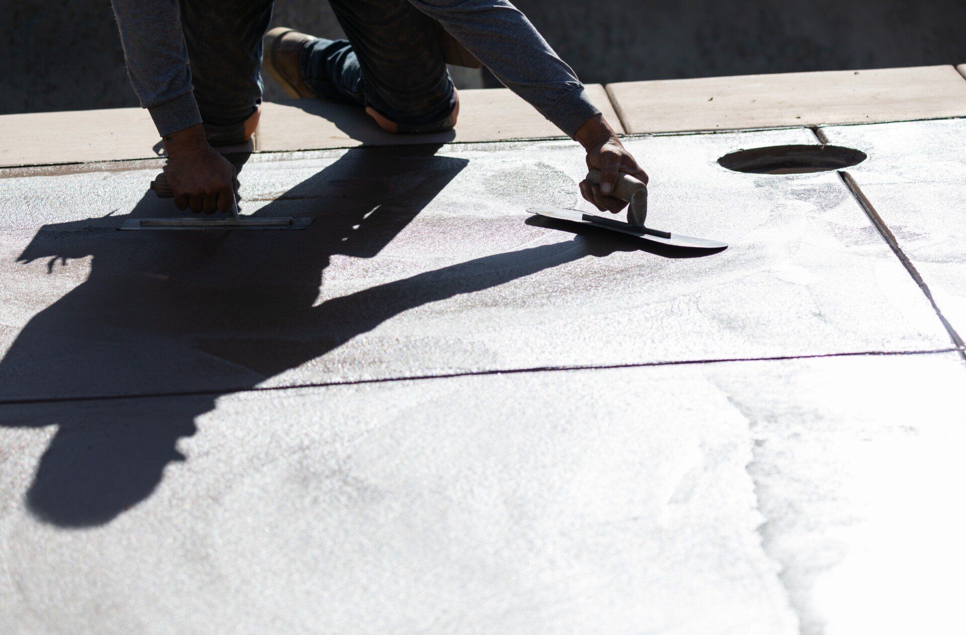 A person smoothing out wet concrete with a trowel on a sunny day. Their shadow is cast on the surface, and nearby, another trowel is visible. The scene suggests construction or repair work by Concrete Contractors specializing in decorative concrete services.